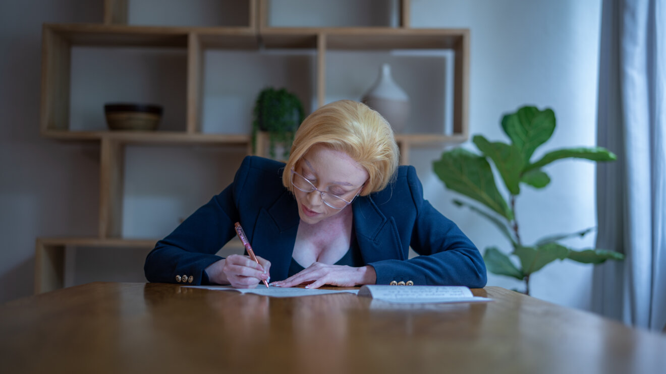 Palesa Mosiea writes a letter at a wooden table in a bright, modern room with plants and shelving in the background, wearing glasses and a navy blazer